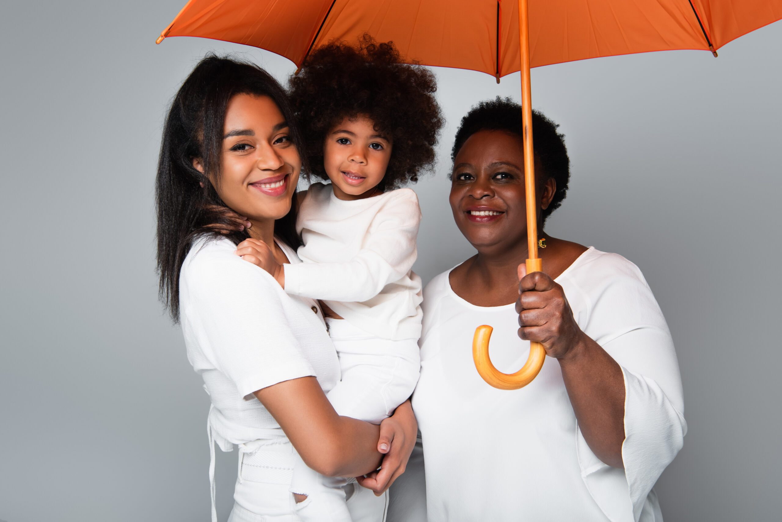 Three generational family standing below an umbrella while smiling at camera. Umbrella insurance.
